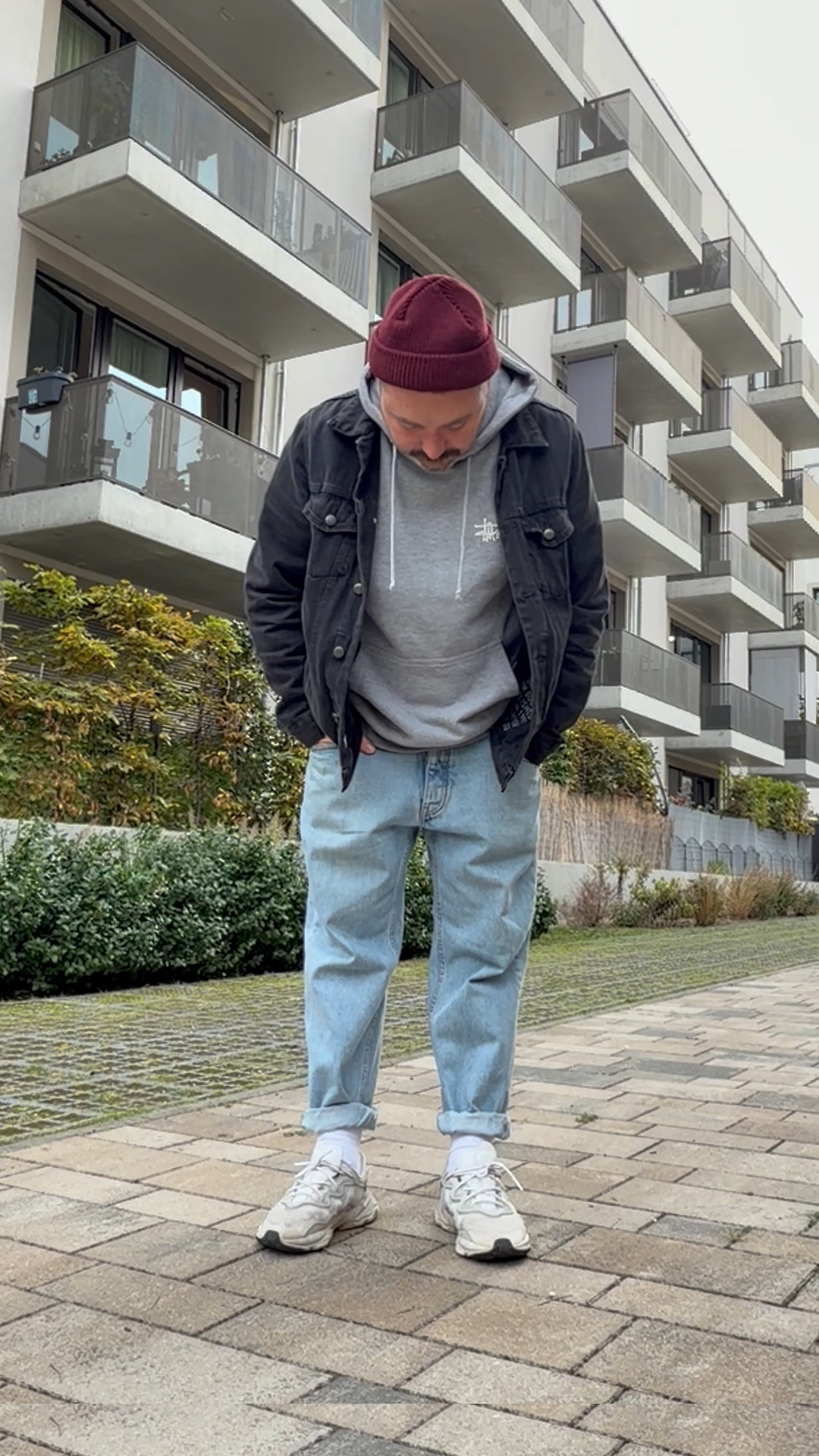 Mann mit bordeauxroter Beanie, grauem Hoodie, schwarzer Jeansjacke und hellblauer Baggy Jeans blickt auf seine weißen Chunky Sneaker.